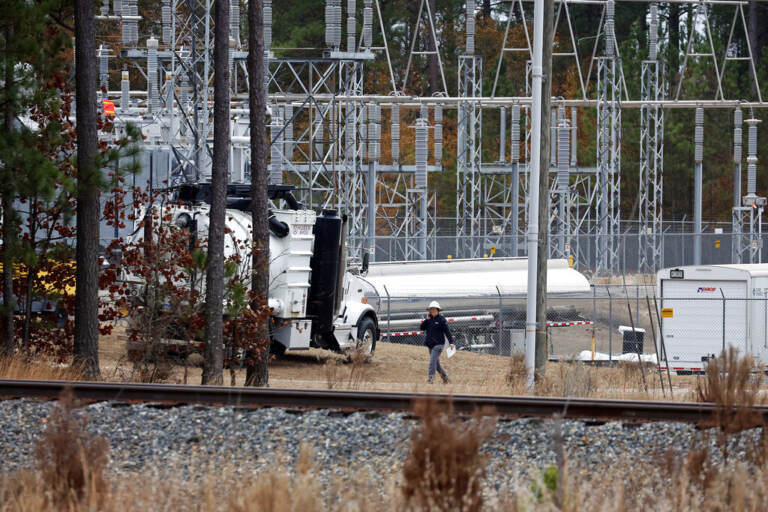 A power substation is shown from a distance. A worker can be seen walking near a tall structure.