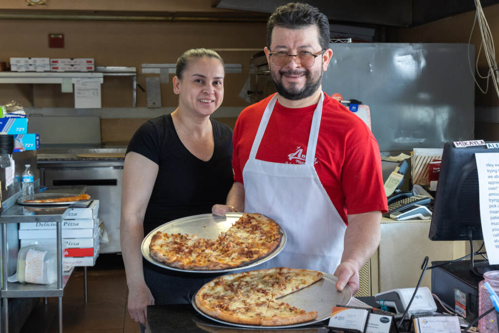Omar Quiñonez and Kelyn Flores pose, smiling, with two pizzas in their kitchen at Amy's Pizzeria.