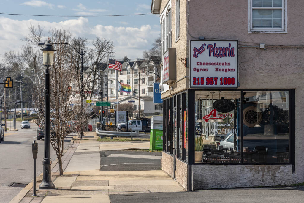 A view from up the street of Amy's Pizzeria. Further down the street are other houses, flags, and cars at an intersection.