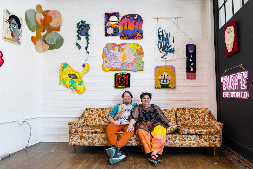 Tim Eads and Tiernan Alexander sit together on a sofa. Tufted artworks are hung on a white wall above their heads.