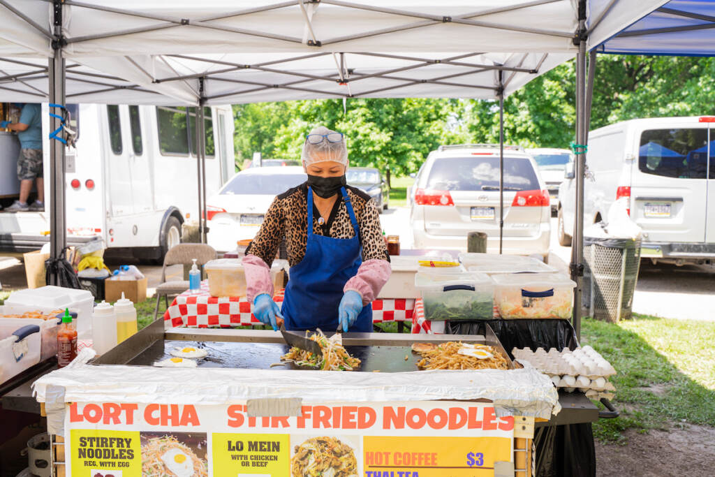 03 29 2023 provided by parks and rec A vendor cooks on a grill at an open stand. Trees and cars are visible behind her.