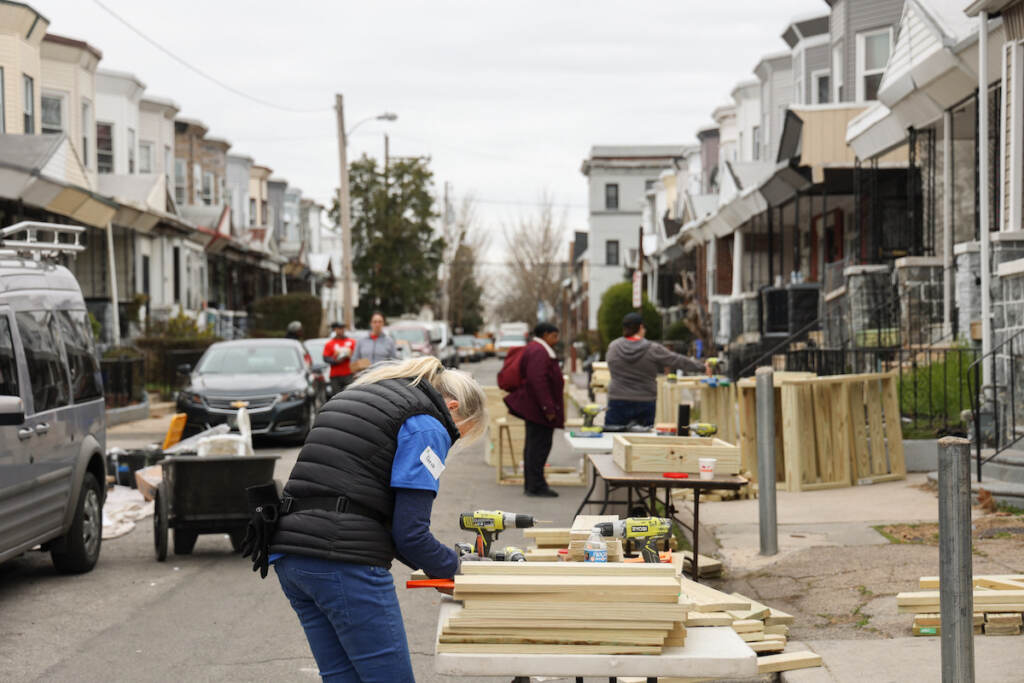 03 31 2023 K paynter rebuilding together philadelphia 5300 block of chancellor street-5 A person works on pieces of wood on a table in the street on a block of rowhomes.