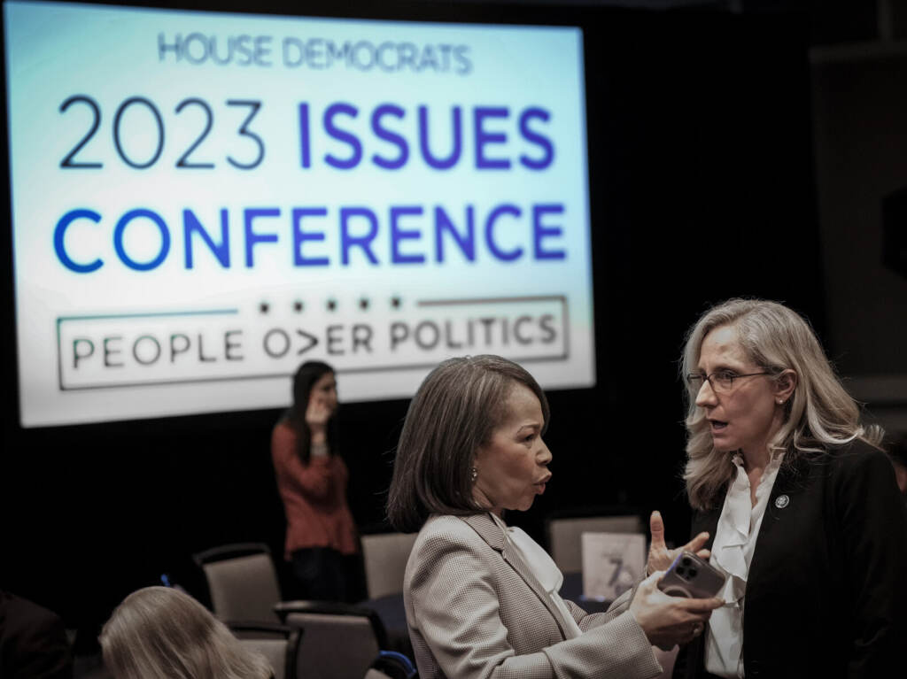 President Biden Speaks At The House Democratic Caucus Issues Conference In Baltimore Two people talk in the foreground. A sign in the background reads: House Democrats 2023 Issues Conference.