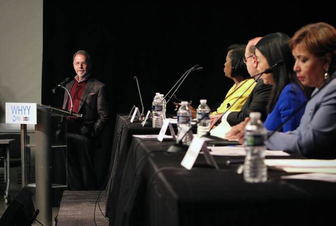 WHYY CEO Bill Marazzo stands by a podium, looking towards Philadelphia mayoral candidates seated in a row at tables onstage.