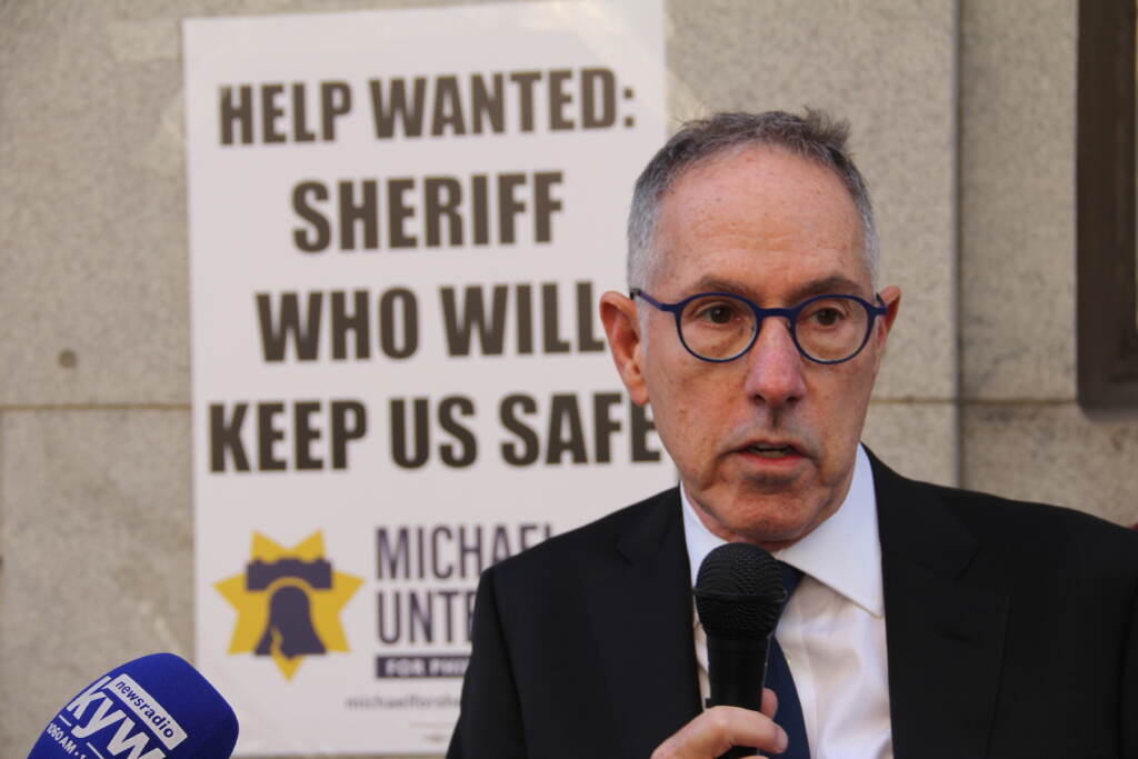 untermeyer-c-sharber-2023-03-07-1 Michael Untermeyer appears with a sign behind him that says ''Help wanted: Sheriff who will keep us safe''