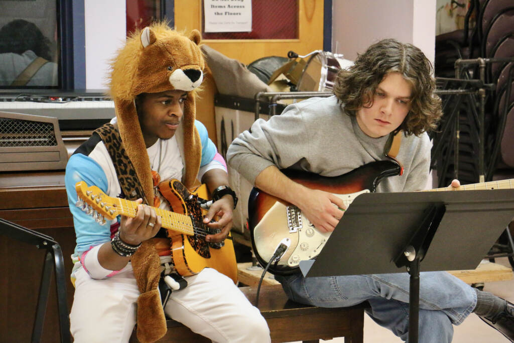 Two students lean together, looking at a piece of sheet music as they play the guitar.