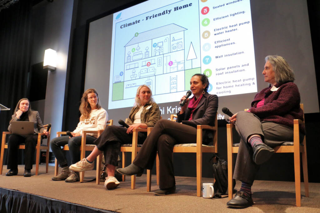 Nidhi Krishen speaks into a microphone while seated onstage as other panelists look on.