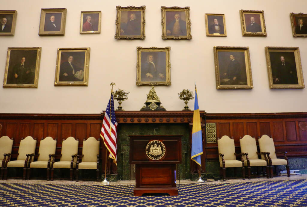2023 04 27-e lee-philadelphia city hall-mayors reception room Portraits of former mayors line the walls of City Hall chambers. A podium and the U.S. flag and the Philadelphia flag are in the center of the photo. Chairs are lined up against the wall.