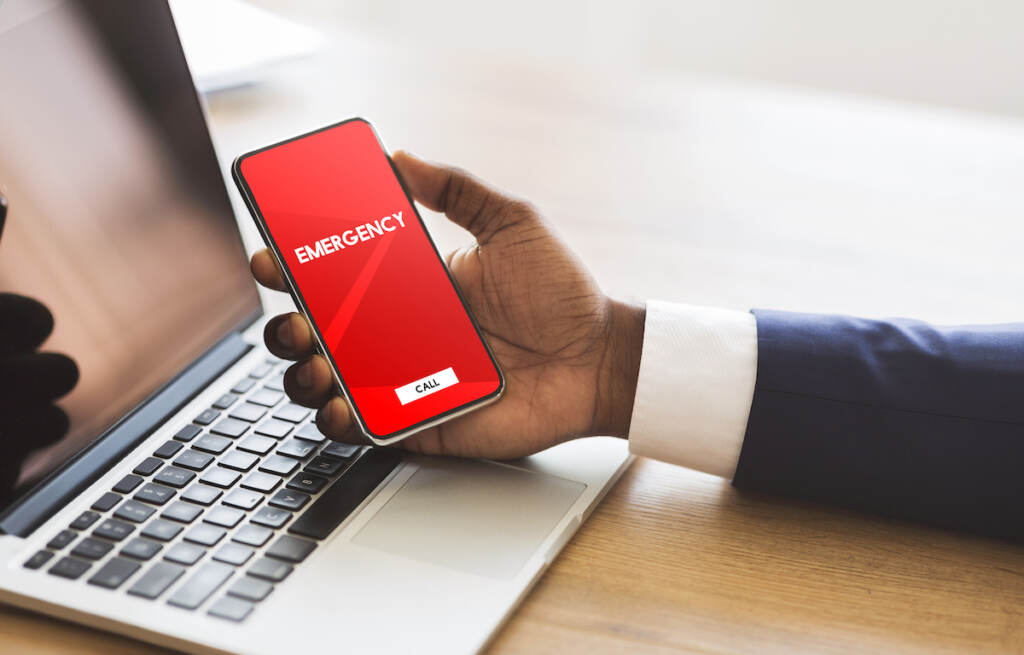 Safety Concept. Male Hand Holding Smart Phone With Emergency Number On Red Screen Over Office Backgr A person holds a phone with a red screen and a box that reads