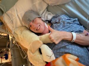 Ashley Yeager lies on a hospital bed with her hand in a thumbs up position.