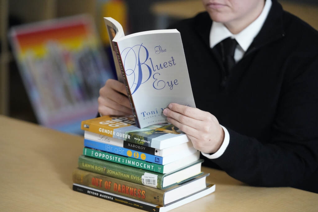 A stack of banned books sits on a table, while someone reads 'The Bluest Eye' by Toni Morrison