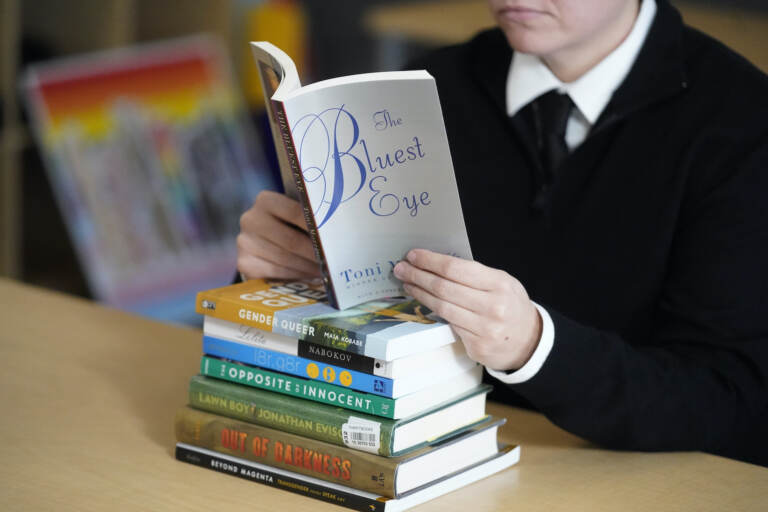 A stack of banned books sits on a table, while someone reads 'The Bluest Eye' by Toni Morrison