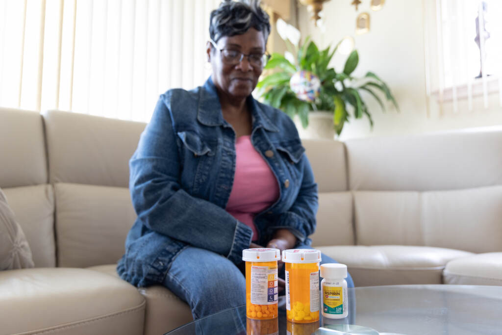 Penny Cooper is seen sitting on a couch, with several medications in the foreground