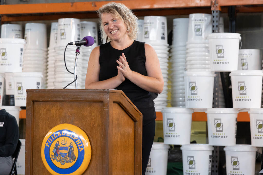 Jen Mastalerz speaks at a podium. Compost bins are visible behind her.