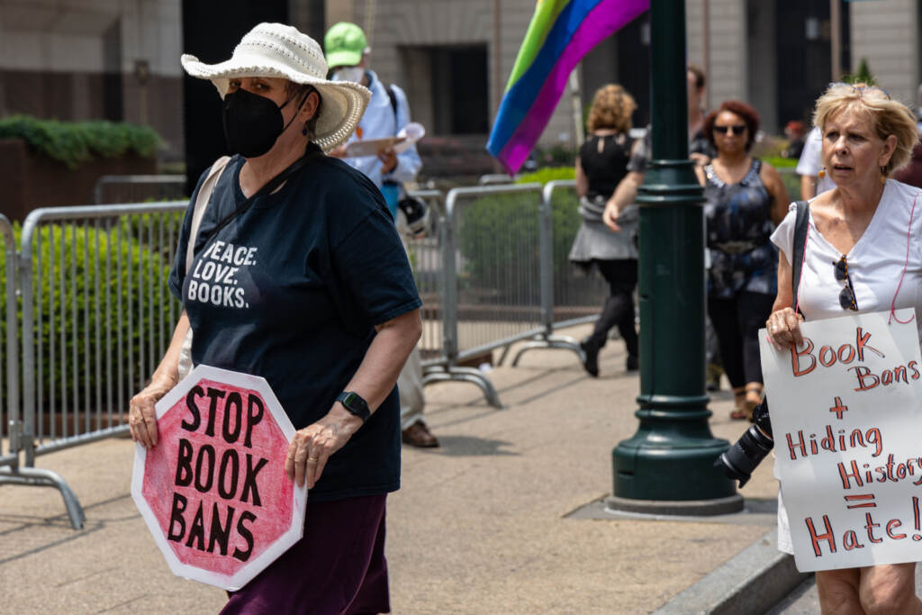 A person wearing a mask protests the Moms for Liberty summit while holding a stop sign-shaped sign that says, ''Stop book bans''