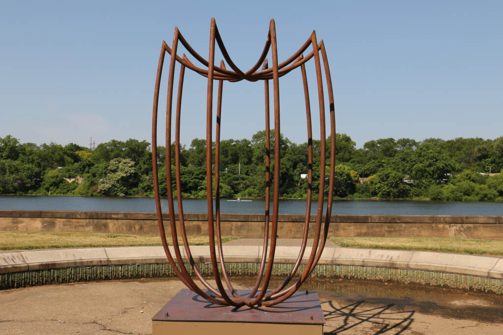 A view of a steel scuplture, behind it are trees and the Schuylkill River, on a sunny day.