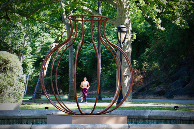 A jogger is visible through the bars of a vessle-shaped steel sculpture.