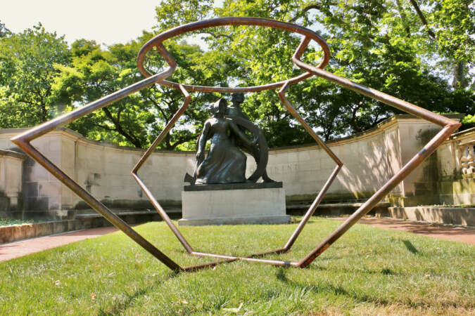 Two sculpted figures are visible through the bars of a steel sculpture in the foreground.