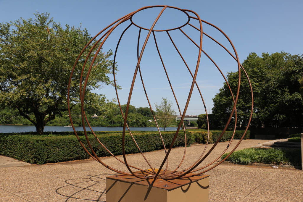 A sculpture made of steel rods forming a sphere is in the foreground. Behind it are trees, the river, and a pathway.