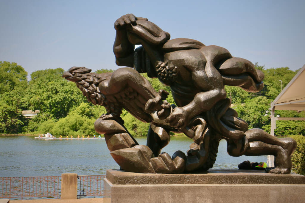 Sculpted figures are visible in the foreground. Behind them is the Schuylkill River, rowers, and trees.