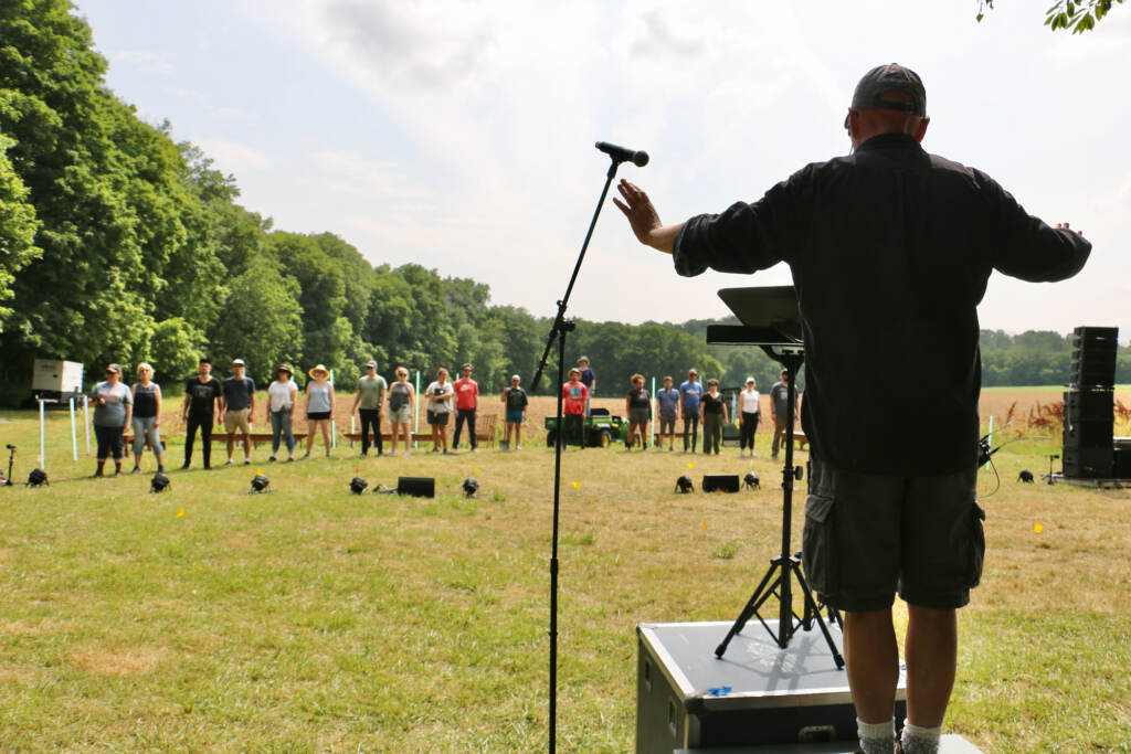 2023 06 20-e lee-donald nally-newtown pa-crossing choir farming conductor Donald Nally stands in front of a group of people, conducting the choir in an open field.