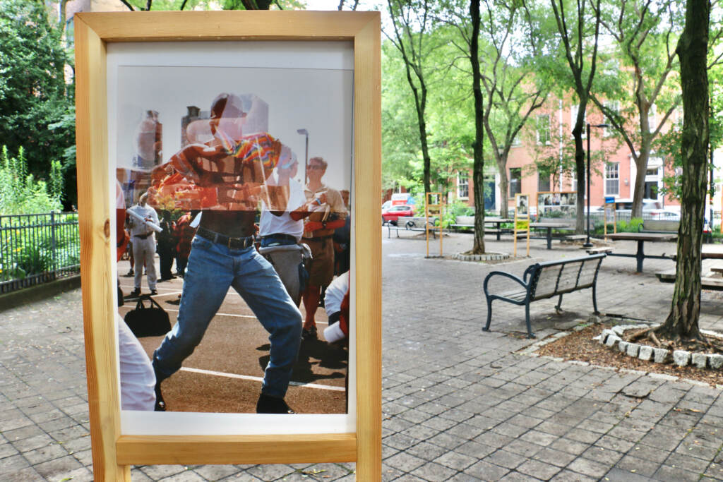 2023 06 27-e lee-philadelphia louis kahn park-and into the streets au courant exhibit double exposure A close-up of a double-exposure photo printed and displayed in a park showing a shirtless man overlayed with a Pride flag.