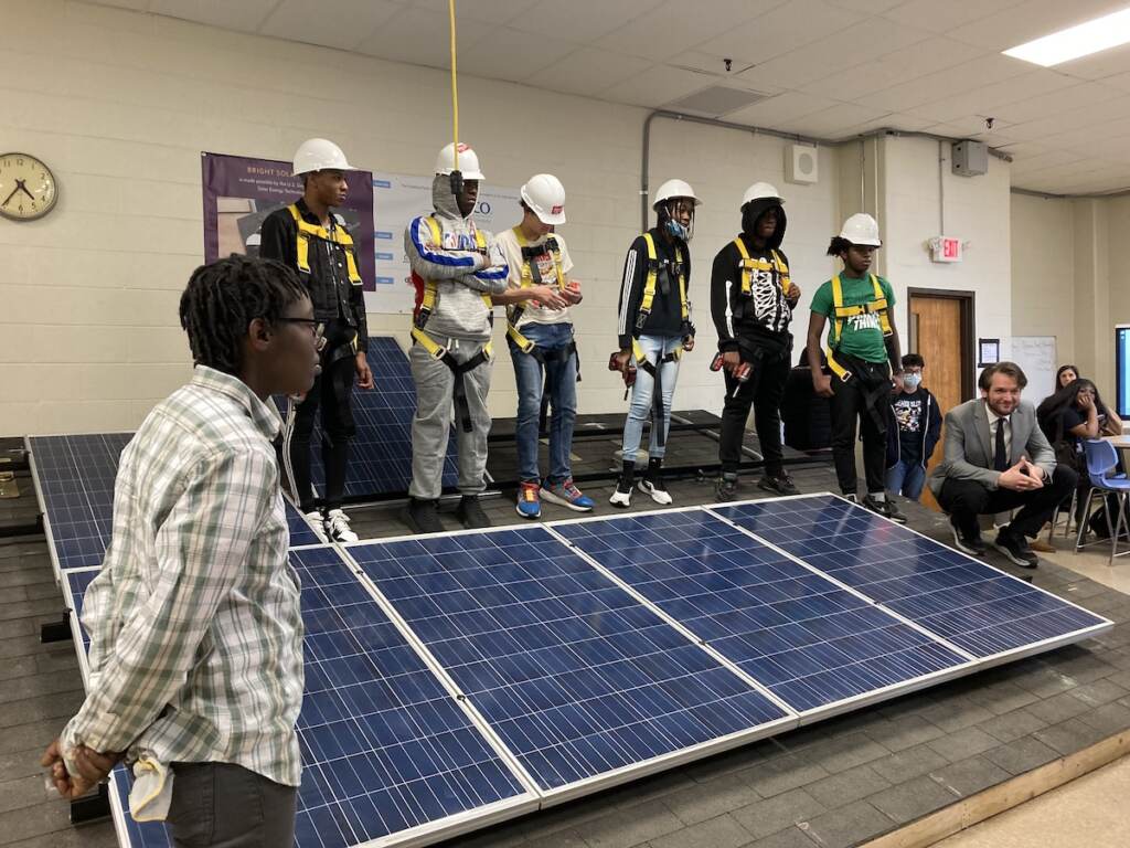 BrightSolarFuturesStudents Anthony Cruz (back row, second student with hard hat from right), stands behind a solar panel with his classmates.