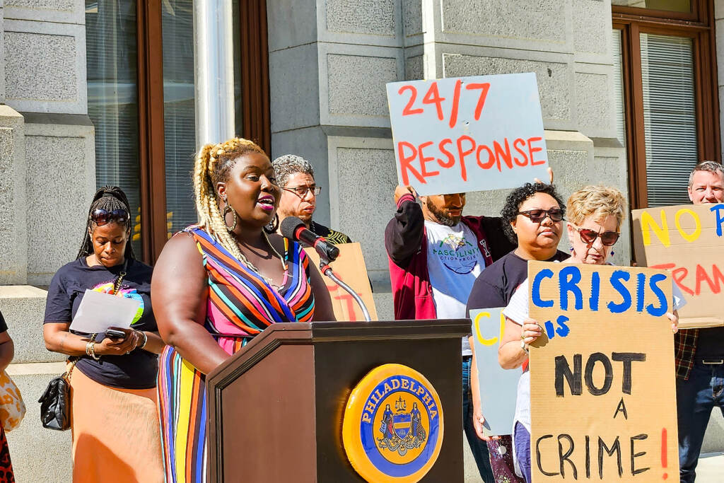 Philly-Treatment-Not-Trauma A woman speaks at a podium, with others holding signs next to her.