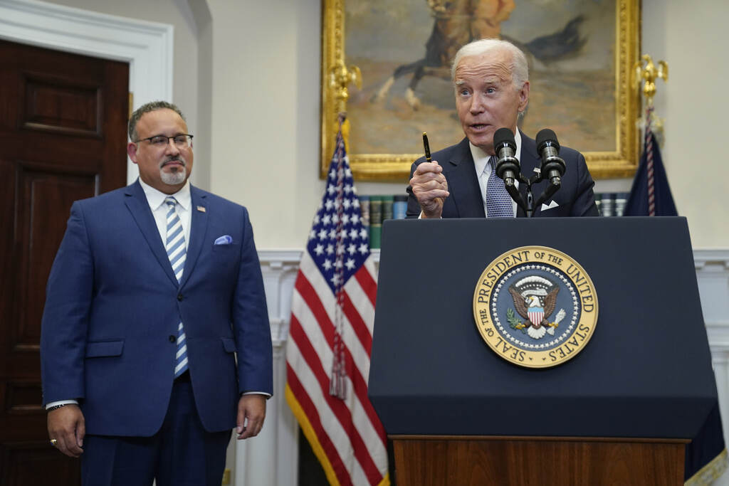 President Joe Biden speaks at a podium. Education Secretary Miguel Cardona listens at left.