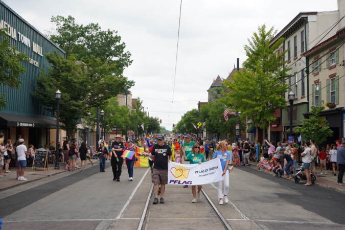 Pride parade participants walk down the street together, carrying Pride flags and other signs.