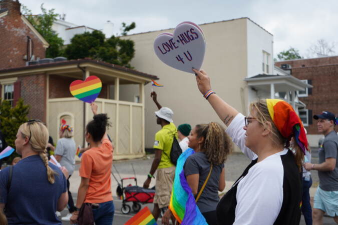 A person holds up a sign that reads Love + Hugs 4 U as they march with a group of people carrying Pride flags.
