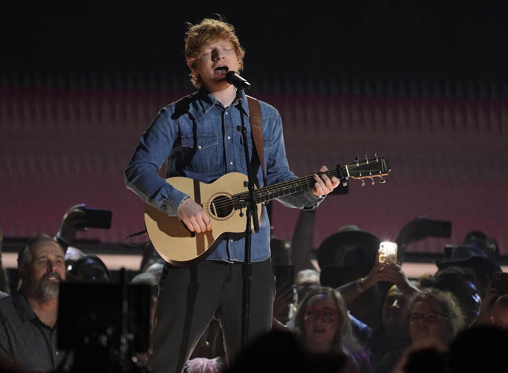 Ed Sheeran sings into a microphone while playing a guitar onstage.