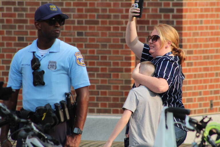 A person covers up a child's face as they walk forward. Next to them a police officer looks on.