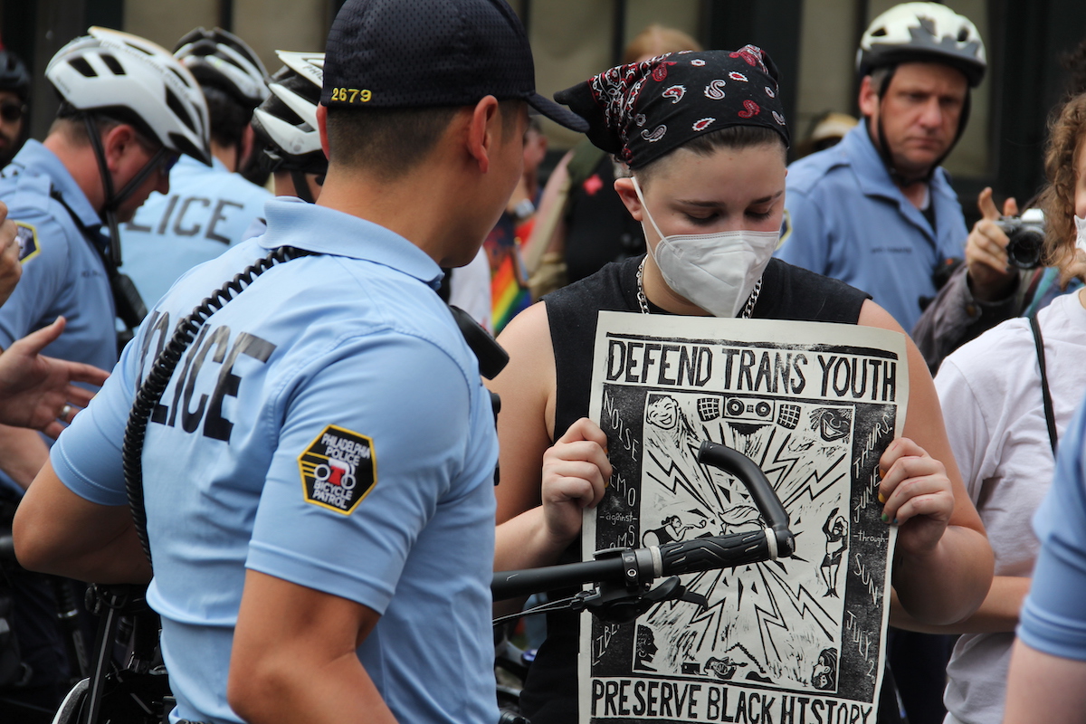A person standing next to police officers holds up a sign that reads Defend Trans Youth, Preserve Black History