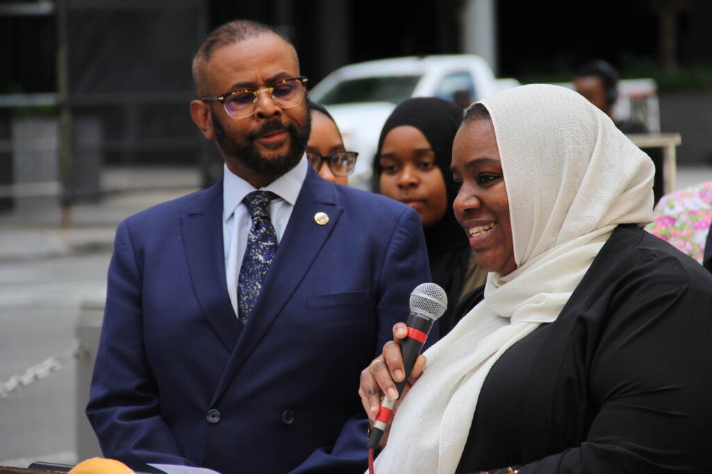 Curtis Jones Jr. looks on as Salima Suswell speaks into a microphone
