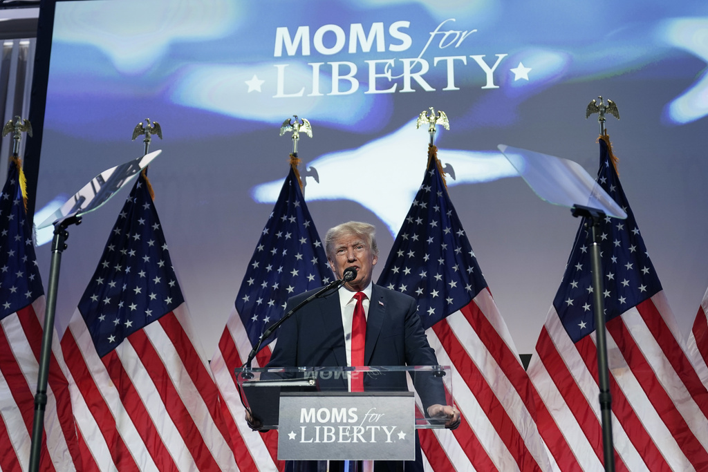 Trump speaks at a podium with a placard that reads Moms for Liberty. Behind him are U.S. flags.