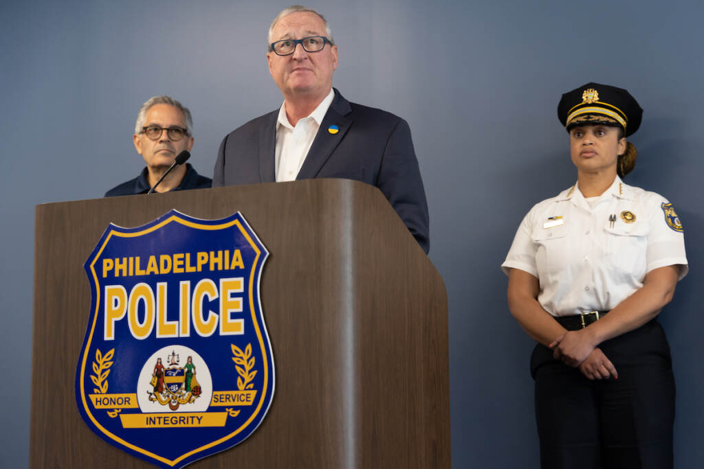 Mayor Kenney speaks at a podium while DA Krasner and Commissioner Outlaw look on.