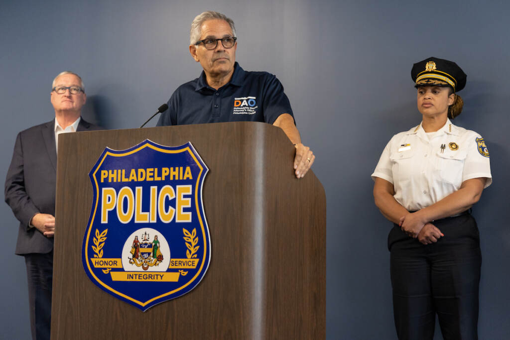 DA Larry Krasner speaks at a podium as Mayor Kenney and PPD Commissioner Outlaw look on.