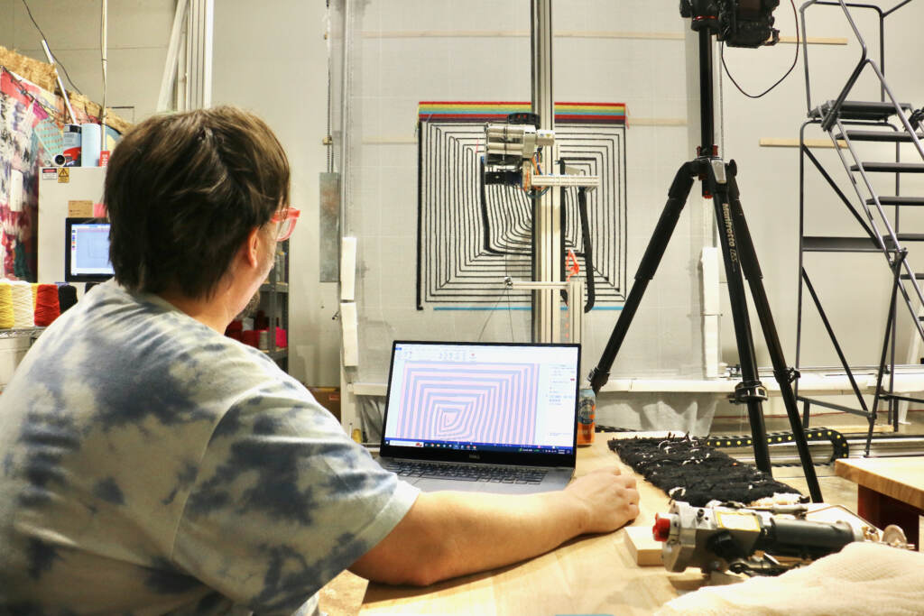 2023 07 07-e lee-tim eads-southwest philadelphia tuft the world-artificial intellignce rugs ai software Tim Eads works on a computer. A rug and a tufter machine working on the rug are visible in the background.