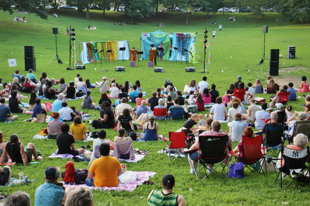 A crowd of people watches performers in the green grass at Clark Park.