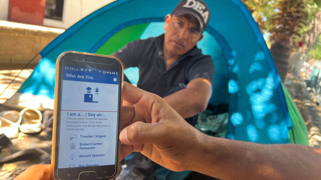 A person holds a phone with a CBP app open on it. Another person looks on in the background.
