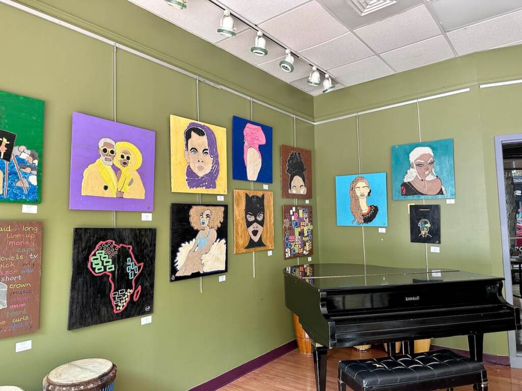 Paintings are hung on a wall in a room. A piano is visible in the corner.