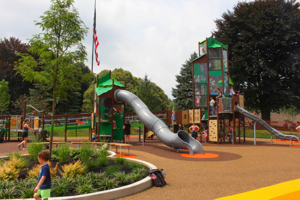 A slide and other structures in the plaground are visible, along with a few children playing, their caregiver, and a U.S. flag waving in the background.