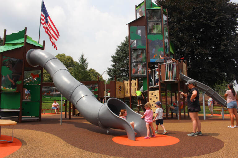 A slide and other structures in the plaground are visible, along with a few children playing, their caregiver, and a U.S. flag waving in the background.