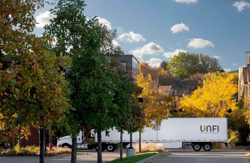 unfi A truck emblazoned with the letters UNFI drives on a road lined with trees.