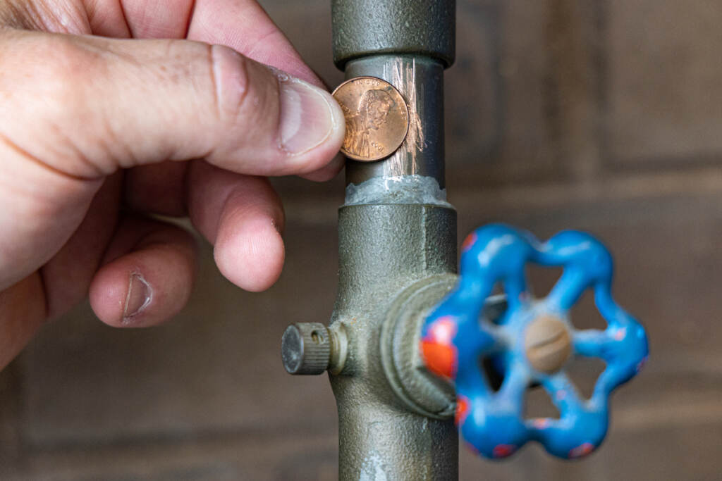 A person holds up a penny next to a scratched pipe, which now matches the color of copper.