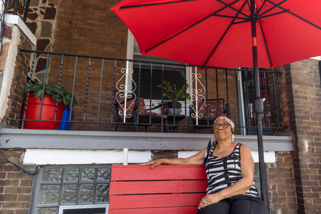 Sharon Drayton sits on a red bench with a red umbrella outside of her home.