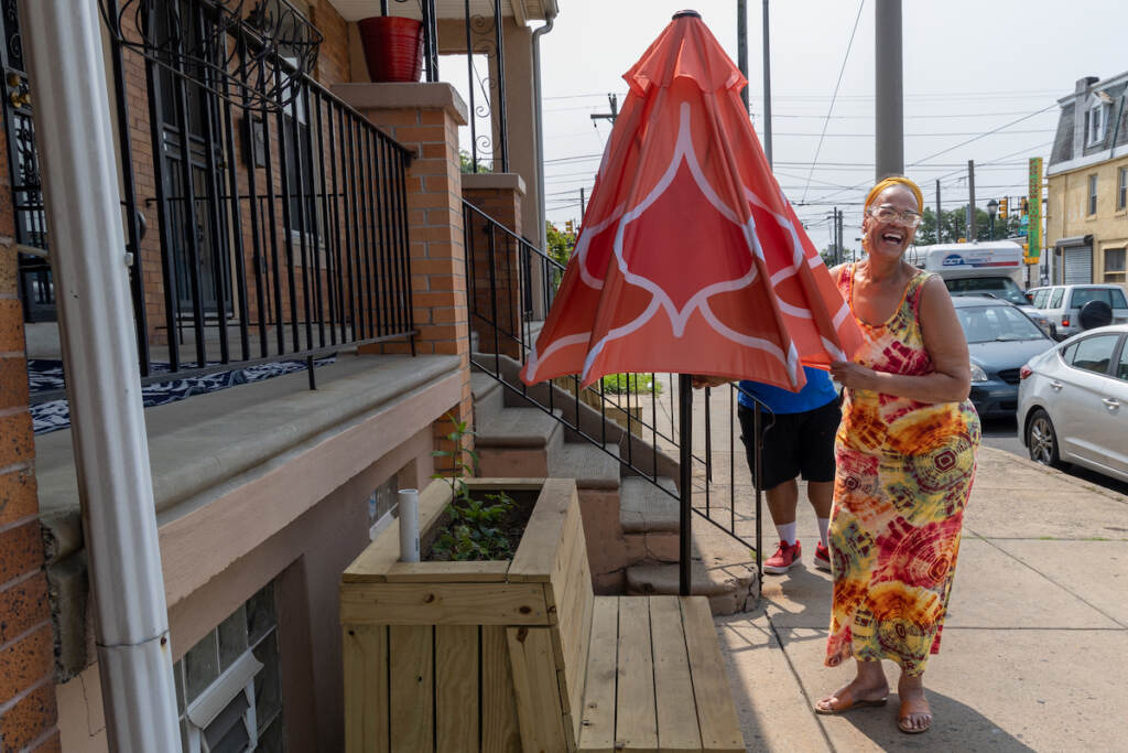 Adrine Wilson smiles and laughs as she stands next to an umbrella on a planter bench outside of a home.
