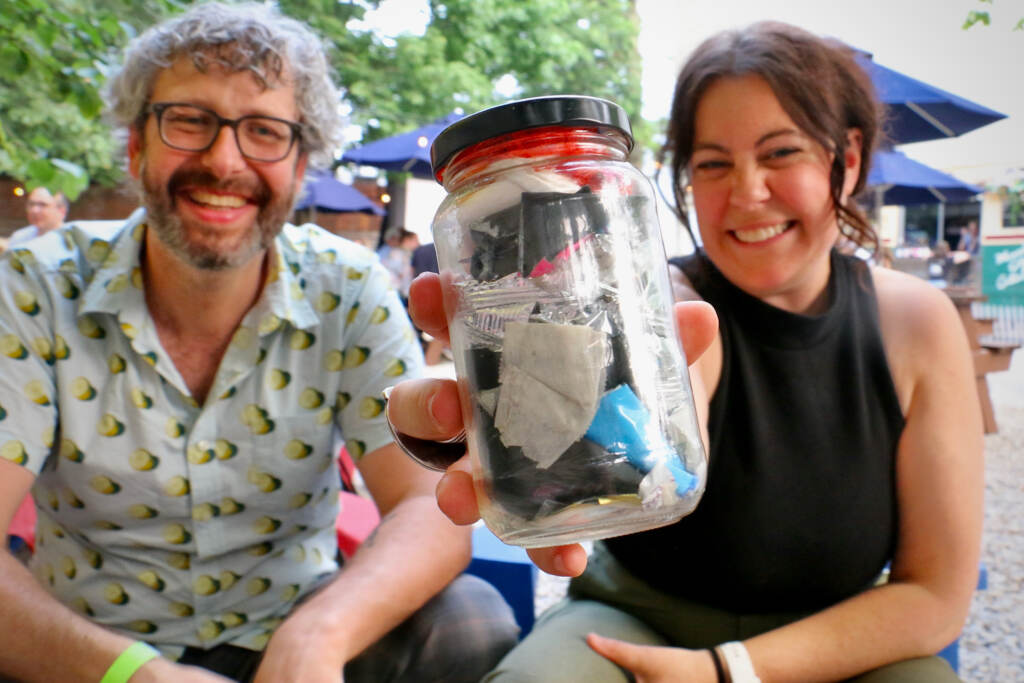 2023 06 29-e lee-austin elston emily gallagher-philadelphia fishtown-zero waste film citywide Emily Gallagher holds up a jar of waste as she smiles, seated next to her husband, Austin Elston, who is also smiling.