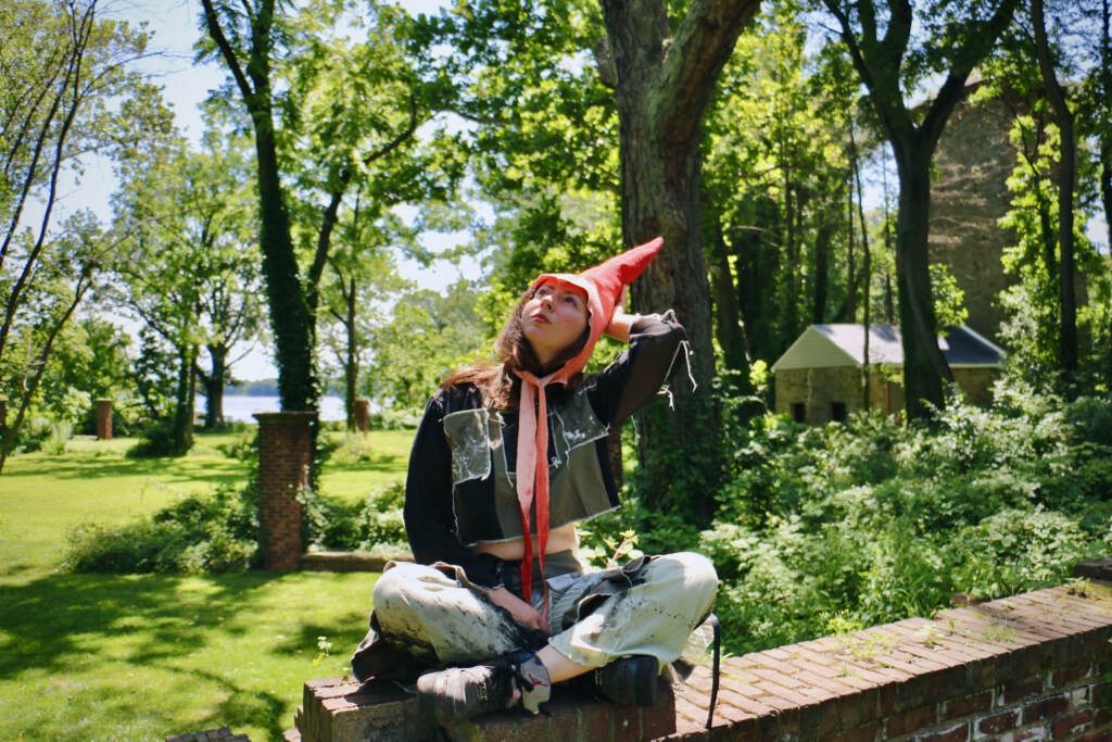 Artist in residence Alex Tatarsky, wearing a gnome costume, looks up towards the sky. Trees and grass are visible in the background.
